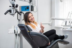 Beautiful female patient with perfect white teeth sitting in dental chair, smiling and showing thumbs up after treatment at modern dental clinic.